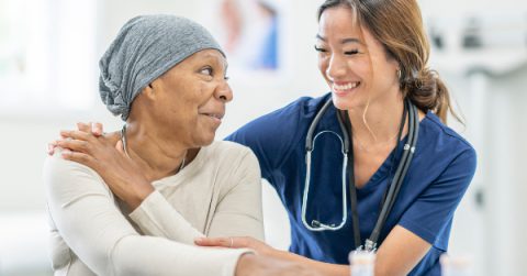 A young nurse of Asian decent bends over beside her patient battling cancer, as she places her hands on her shoulders to comfort her and show support. The patient is dressed comfortably and has a headscarf on to keep her warm as she smiles with the news that her treatments are done.