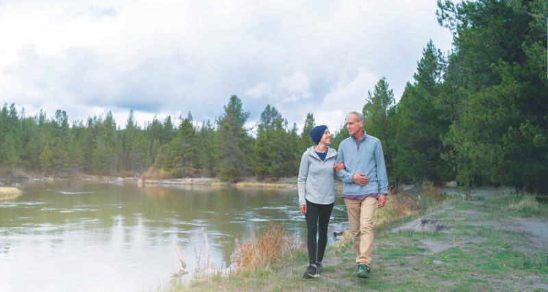 A mixed race millennial age woman with cancer walks next to a river in a forested area with her father. Their arms are linked as they talk and enjoy time together in nature. The woman is looking up at her dad with love and appreciation for his support.