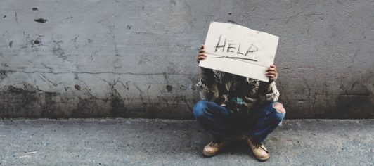 A homeless, long-haired Asian man sits hopelessly leaning against a wall as there is no one to help him with work and food in his hand holding a sign for help. Homeless sleep on streets