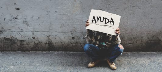 A homeless, long-haired Asian man sits hopelessly leaning against a wall as there is no one to help him with work and food in his hand holding a sign for help. Homeless sleep on streets
