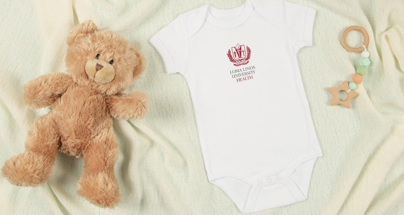 White cotton baby short sleeve bodysuit, teddy bear and natural wooden toy on beige blanket throw background. Infant onesie mockup. Blank gender neutral newborn bodysuit mock up template. Top view.