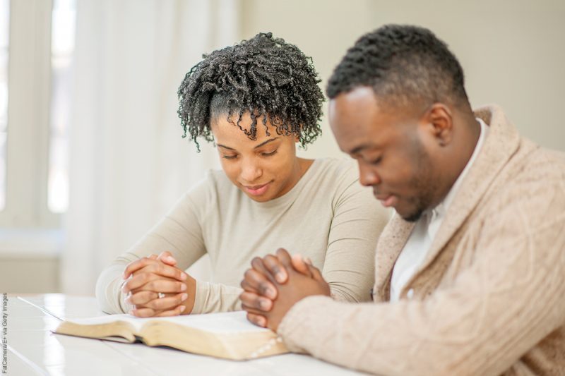 A husband and wife are sitting together at their kitchen table and are praying together.