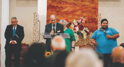 El pastor Peter Neri lee una promesa de la Biblia mientras el pastor Rodolfo Paredes la traduce al español. También en la plataforma, de izquierda a derecha, el pastor Ramón Verduzco y la pastora Melody Darrow.