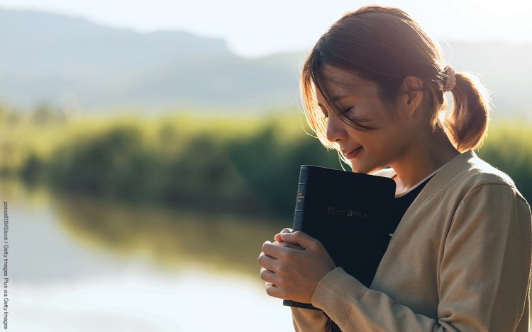 Christian woman praying on the holy bible in a field during a beautiful sunset, holding the bible in her hands. The concept of faith, spirituality, and religion. Peace, hope. Wishing for a better life