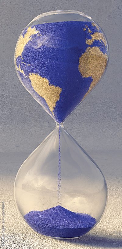 A close up image of an hourglass filled with blue and yellow sand, in the pattern of the countries of the earth. Sand is slowly trickling from the top to pile up in the bottom chamber over time. The timer sits on a rough textured concrete floor near wall.