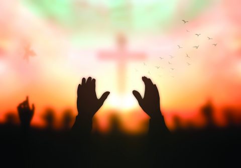 Silhouette christian hand rising over blurred cross on spiritual light background