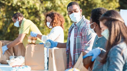 A mid adult man places a canned food item into a paper bag as he volunteers with his teenage son at an outdoor food bank. He and other volunteers are wearing protective face masks as they are volunteering during the coronavirus pandemic.