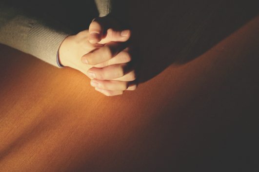 The sun shining on a manâs praying hands on a wooden table, prayer of obedience