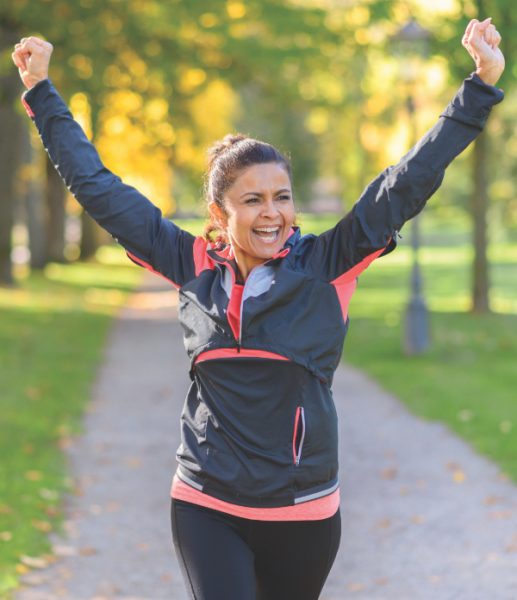 Happy fit middle aged woman cheering and celebrating as she walks along a rural lane through a leafy green park after working out jogging