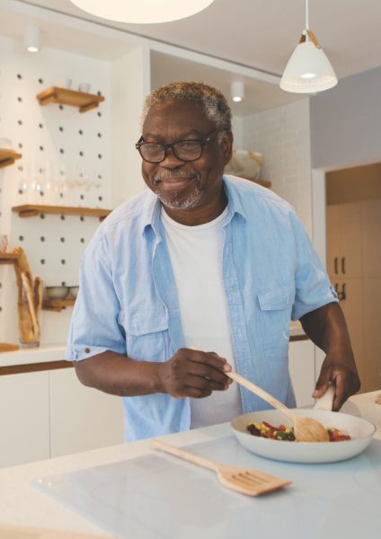 Senior African man standing in the kitchen, watching recipe on laptop and preparing a healthy meal.