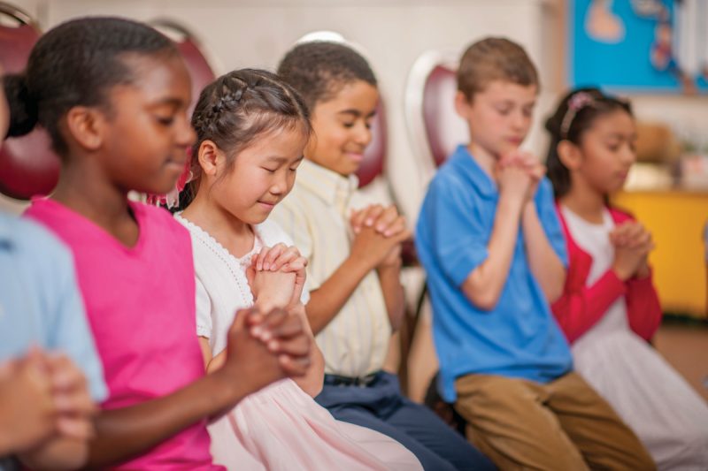 A multi-ethnic group of elementary age children are kneeling on the floor and are praying together.