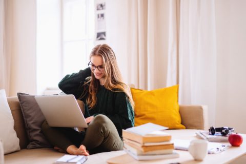 A young happy college female student sitting on sofa at home, using laptop when studying.