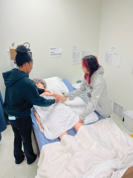 Las estudiantes de último año Berenice Rwamatwara (izquierda) y Kaitlyn Ordas (derecha) practican rodar a un paciente sobre su costado usando el maniquí de CNA.